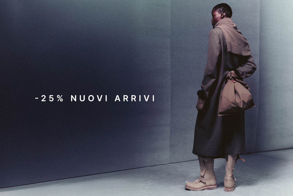 Woman in long dark coat and boots holding beige handbag against minimalist grey backdrop, text '-25% nuovi arrivi' (25% off new arrivals)
