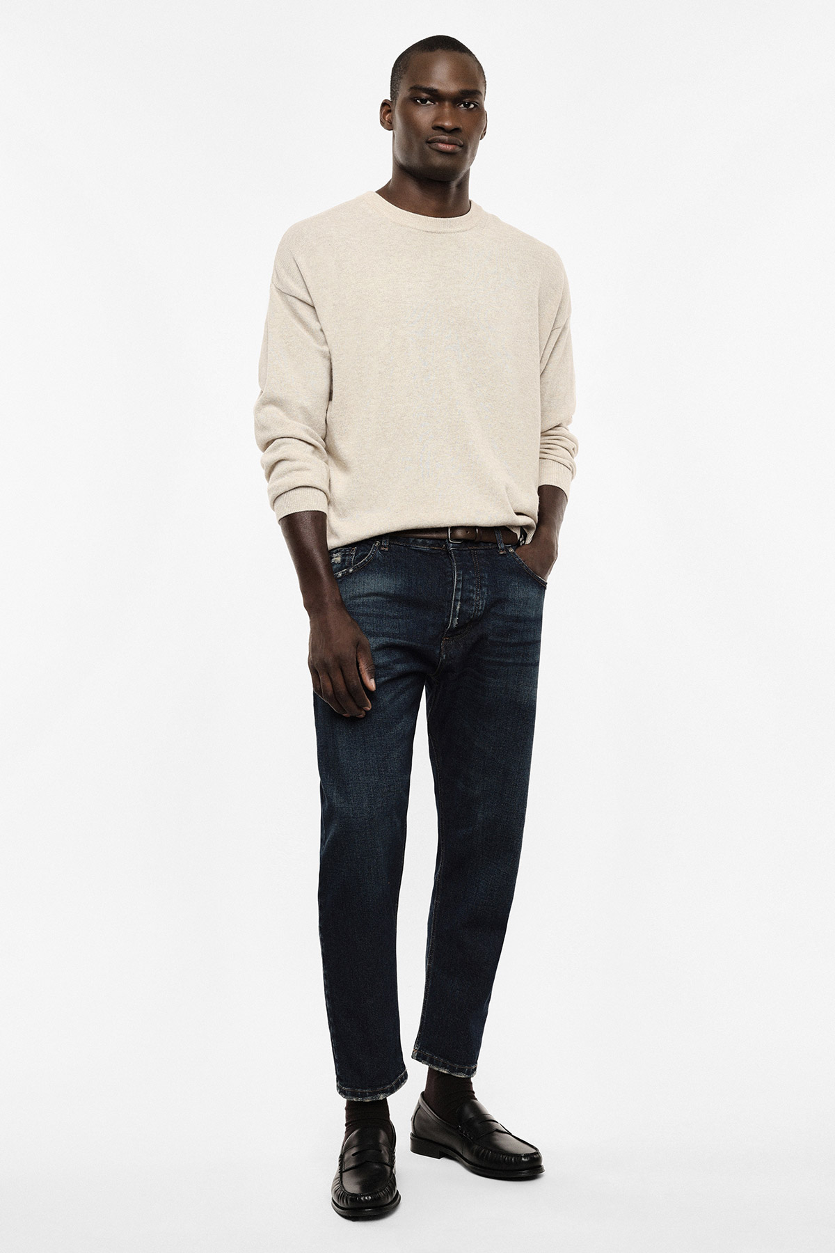 Male model wearing a beige crew-neck sweater, dark blue jeans and black loafers, standing with hands in pockets on white background