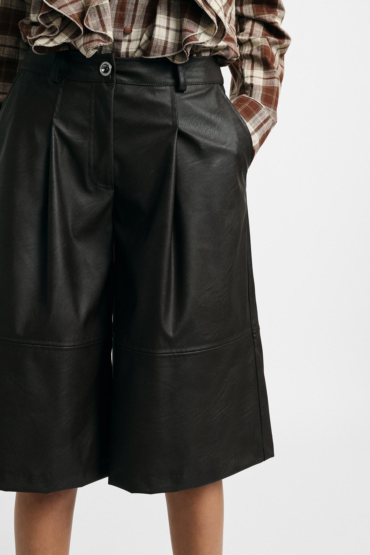 Model wearing black faux-leather culotte shorts with pleats and a brown checked shirt, hands in pockets