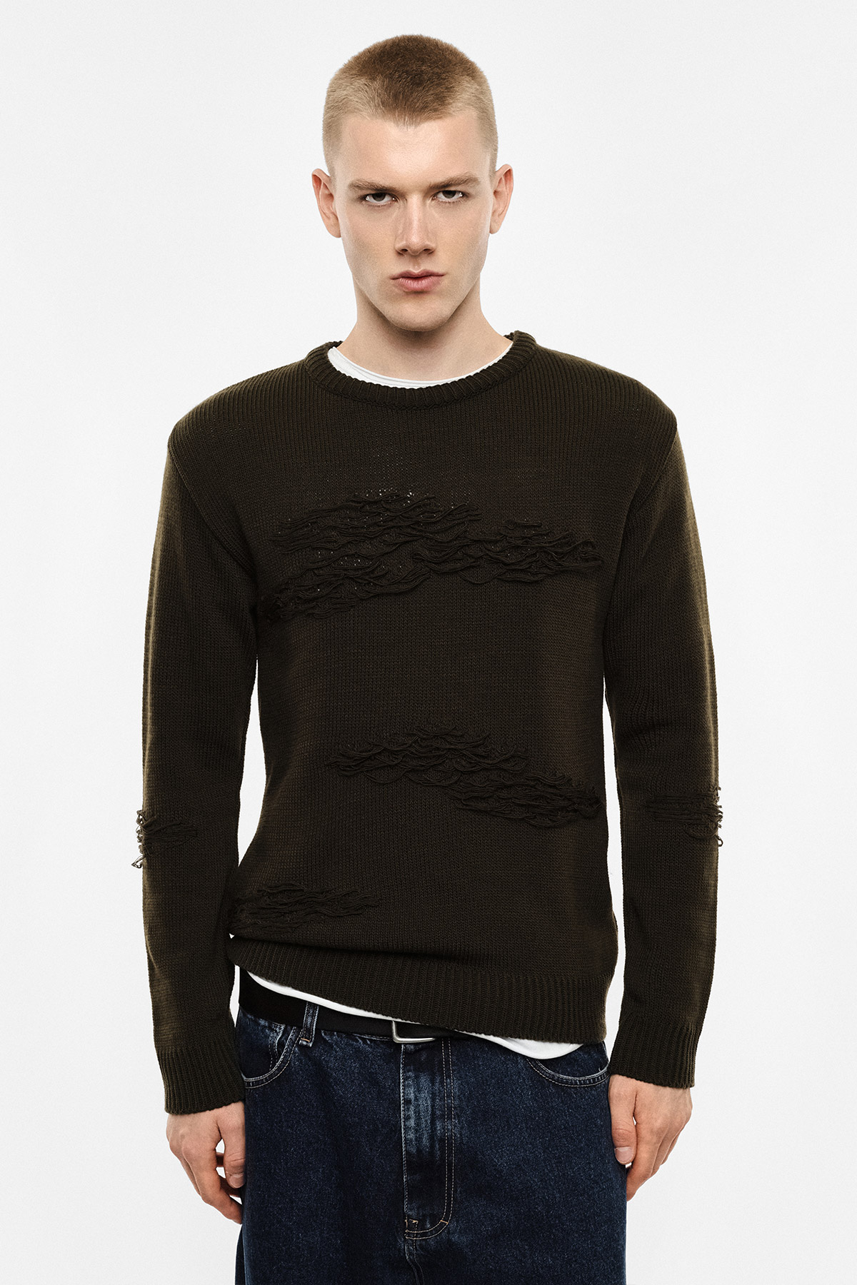 Male model wearing a dark olive distressed crew‑neck sweater and blue jeans, standing front‑on against a plain white studio background