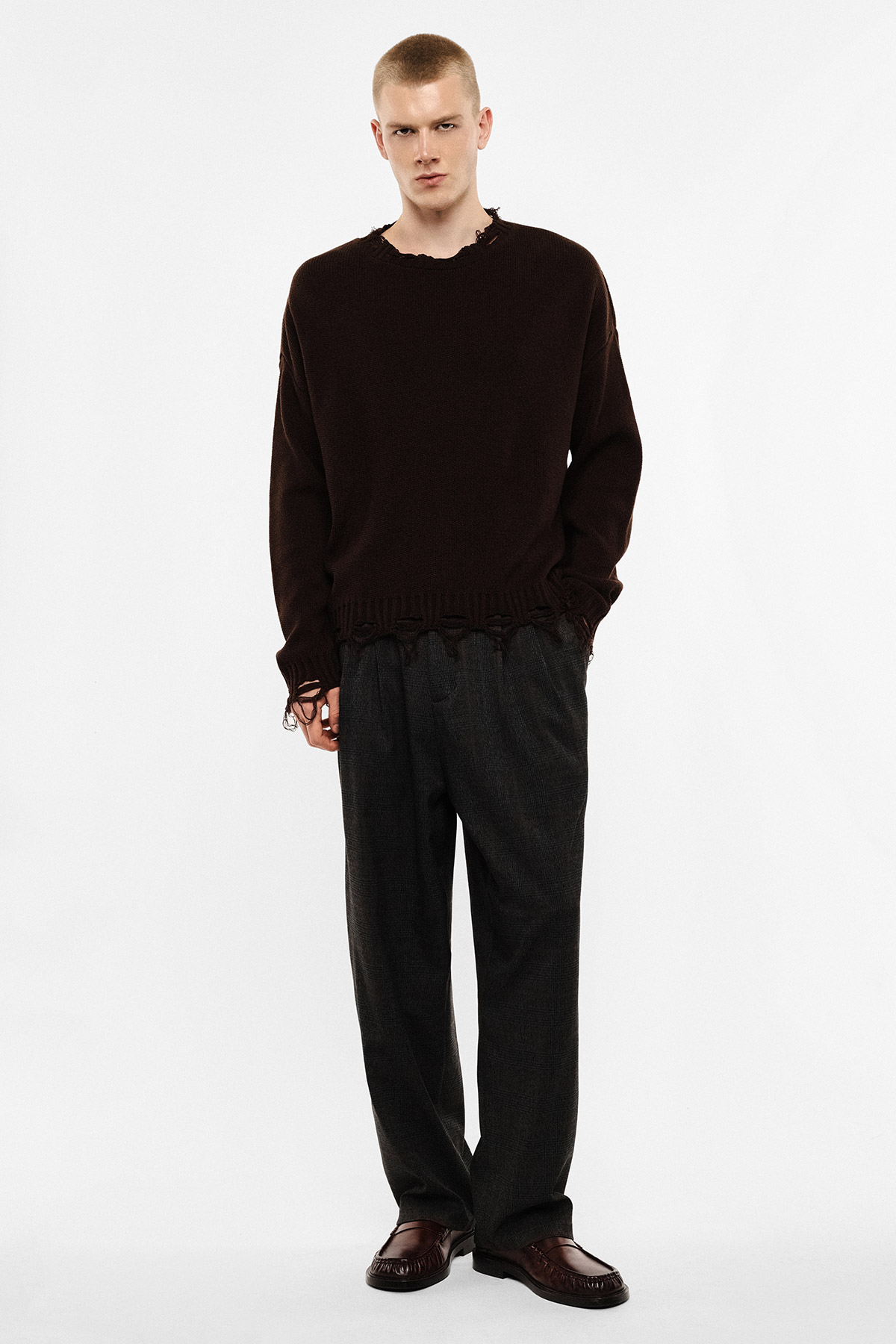 Full-length male model in dark brown distressed knit sweater, grey wide leg trousers and brown loafers on white background