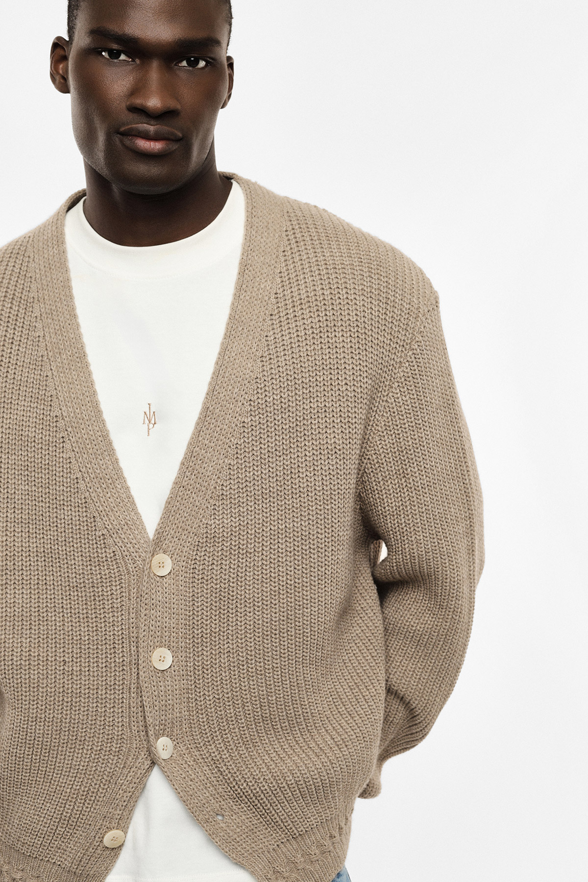 Male model wearing a beige chunky knitted cardigan over a white T‑shirt, close-up studio shot
