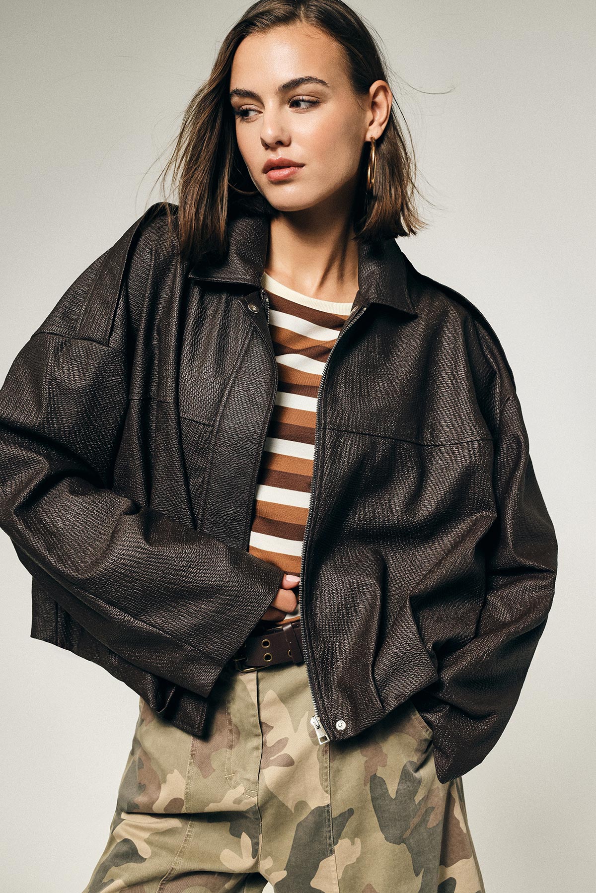 Fashion model wearing oversized brown textured leather jacket over brown-and-white striped top and camouflage trousers, looking to the right
