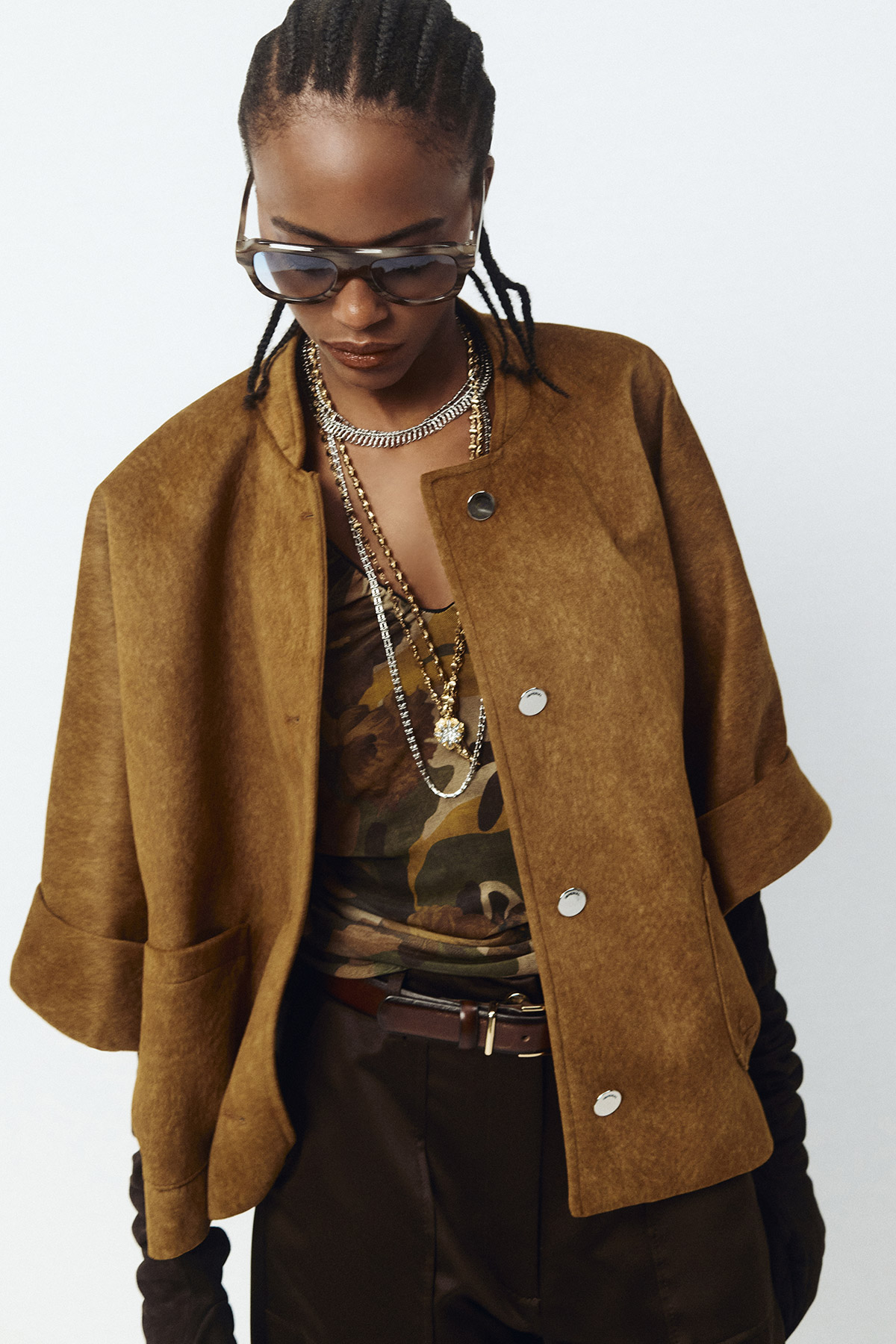 Fashion portrait: person wearing brown suede jacket, camouflage top, layered gold necklaces, sunglasses and leather gloves