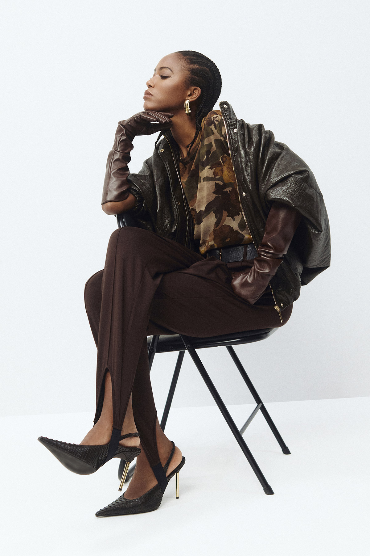 Seated woman with braided hair wearing camo top, oversized leather jacket, long brown gloves, split-hem trousers and pointed heels