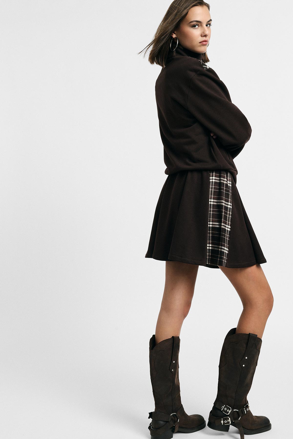Woman modelling brown jacket and flared skirt with plaid panel, wearing knee-high brown boots, standing sideways against pale background