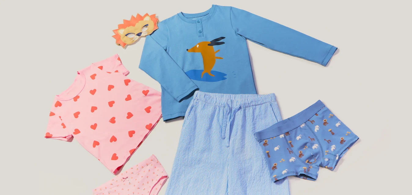 Pigiama e biancheria intima per bambini in rosa e blu con motivi giocosi, oltre a una piccola maschera da leone, disposti su uno sfondo semplice.