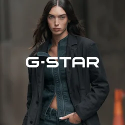 Donna con capelli lunghi e scuri che indossa una giacca scura aperta sopra un denim aderente e un blazer scuro, in piedi in un ambiente urbano.