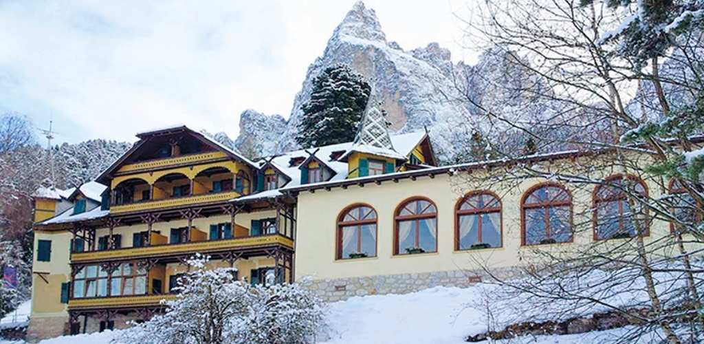 Yellow alpine hotel with wooden balconies and arched windows amid snow, pine trees and a jagged mountain peak in the background