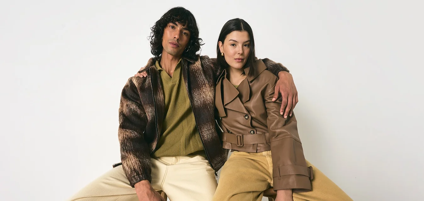Uomo e donna seduti su sgabelli, indossando abbigliamento casual in tonalità terrose, con un braccio attorno l'altro, su uno sfondo bianco.