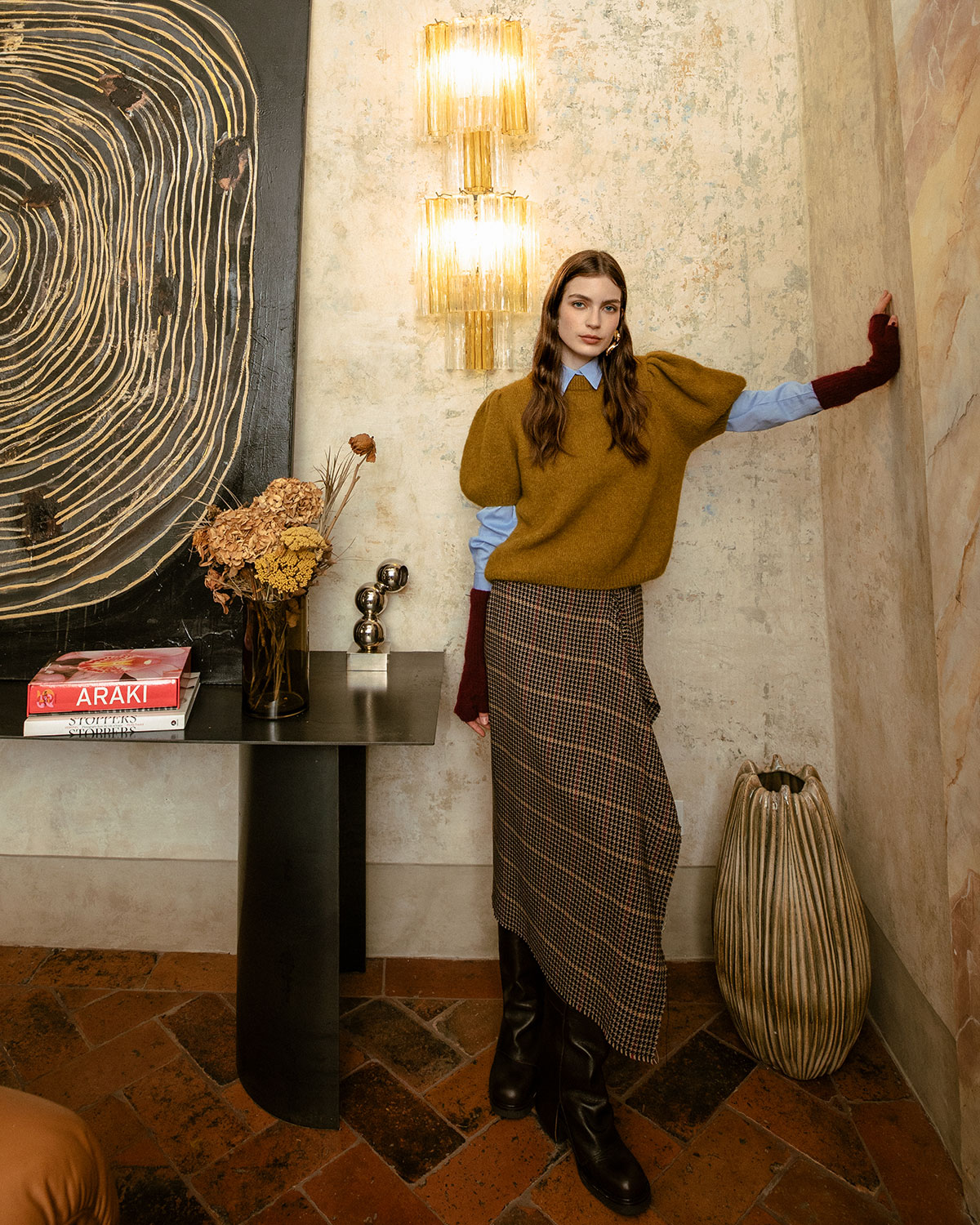 Woman posing against a textured wall in a stylish interior wearing a mustard jumper, blue shirt, houndstooth skirt and black boots beside a console