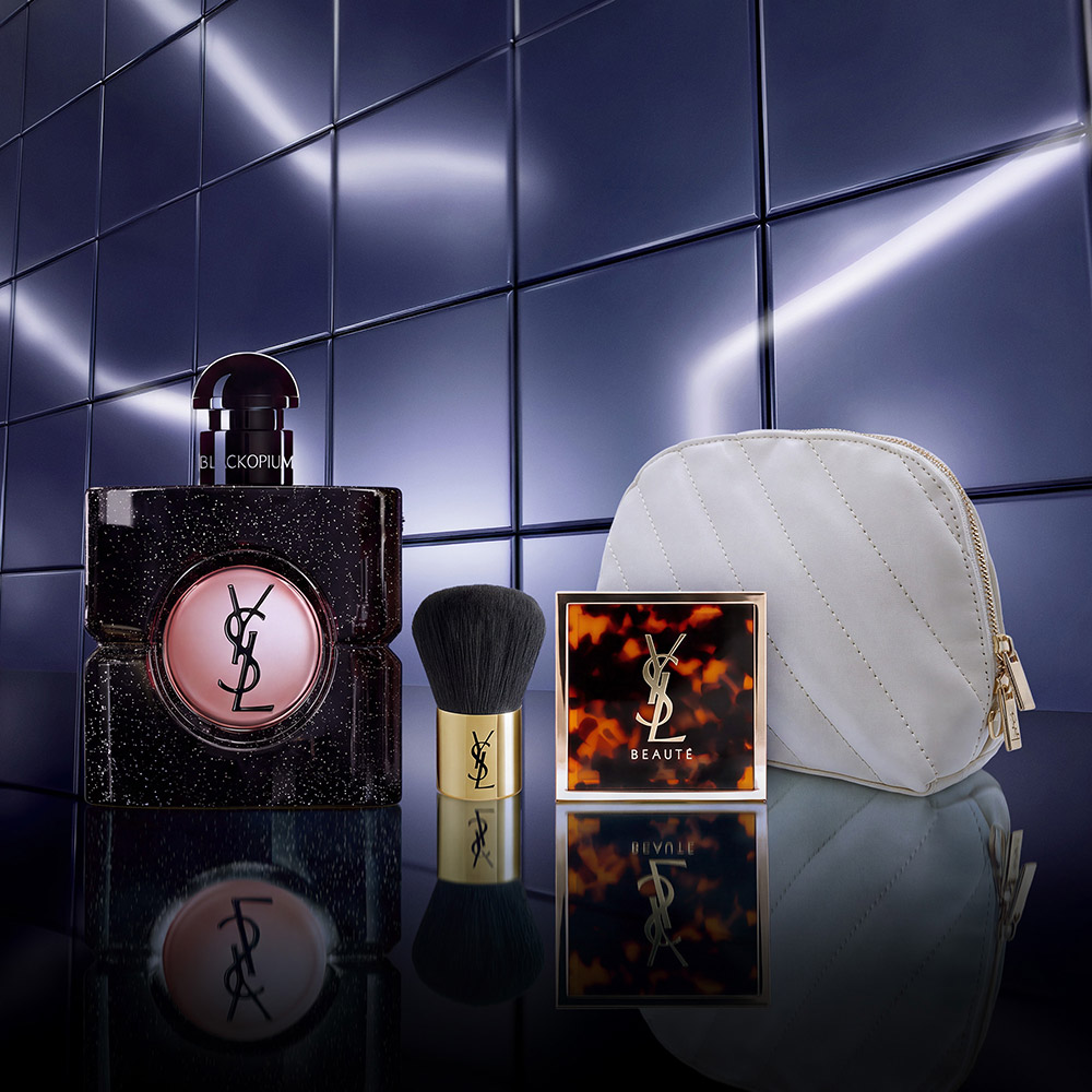 Una trousse bianca trapuntata, un pennello kabuki nero e una cipria tartarugata YSL su sfondo di piastrelle viola.