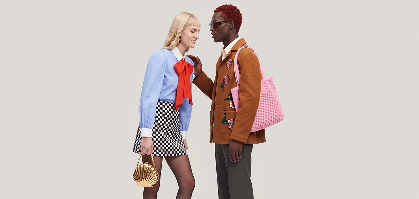 Due persone vestite con stile stanno una di fronte all'altra, una con una piccola borsa dorata e l'altra con una borsa tote rosa.