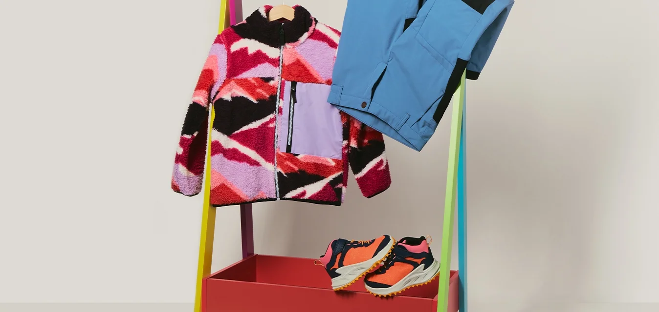 Giacca in pile colorata, pantaloni blu appesi a un supporto multicolore con scarpe da trail running arancioni e nere sotto, su uno sfondo grigio.