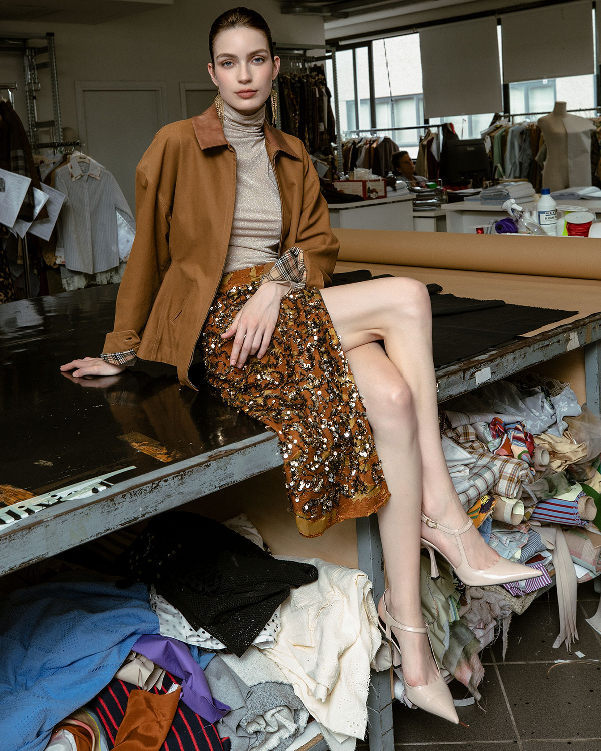 Woman seated on worktable in fashion studio wearing brown jacket, sequinned orange skirt and nude heels amid fabric scraps