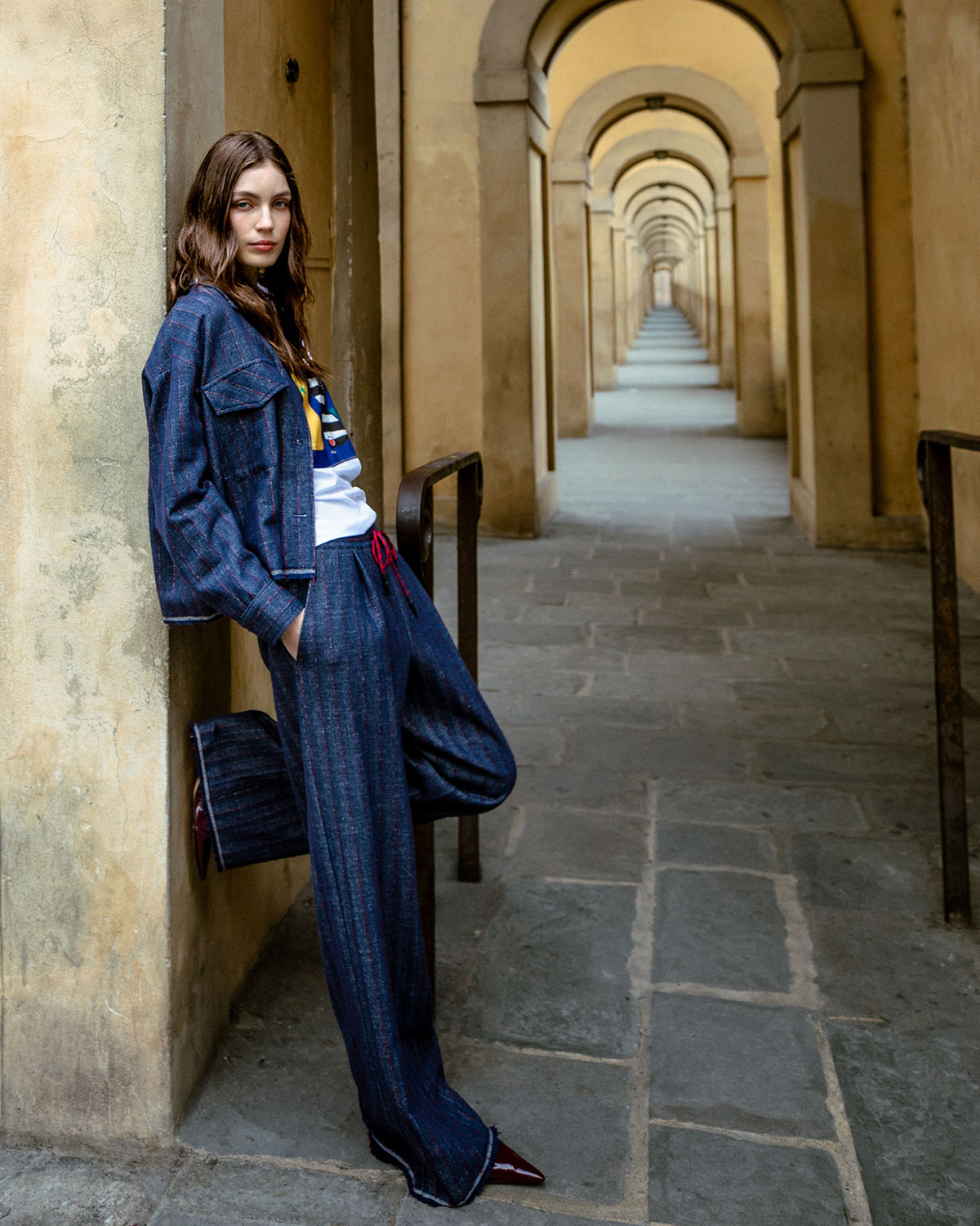 Woman leaning on a column in a stone colonnade, wearing navy pinstripe jacket and trousers, white T-shirt and red shoes