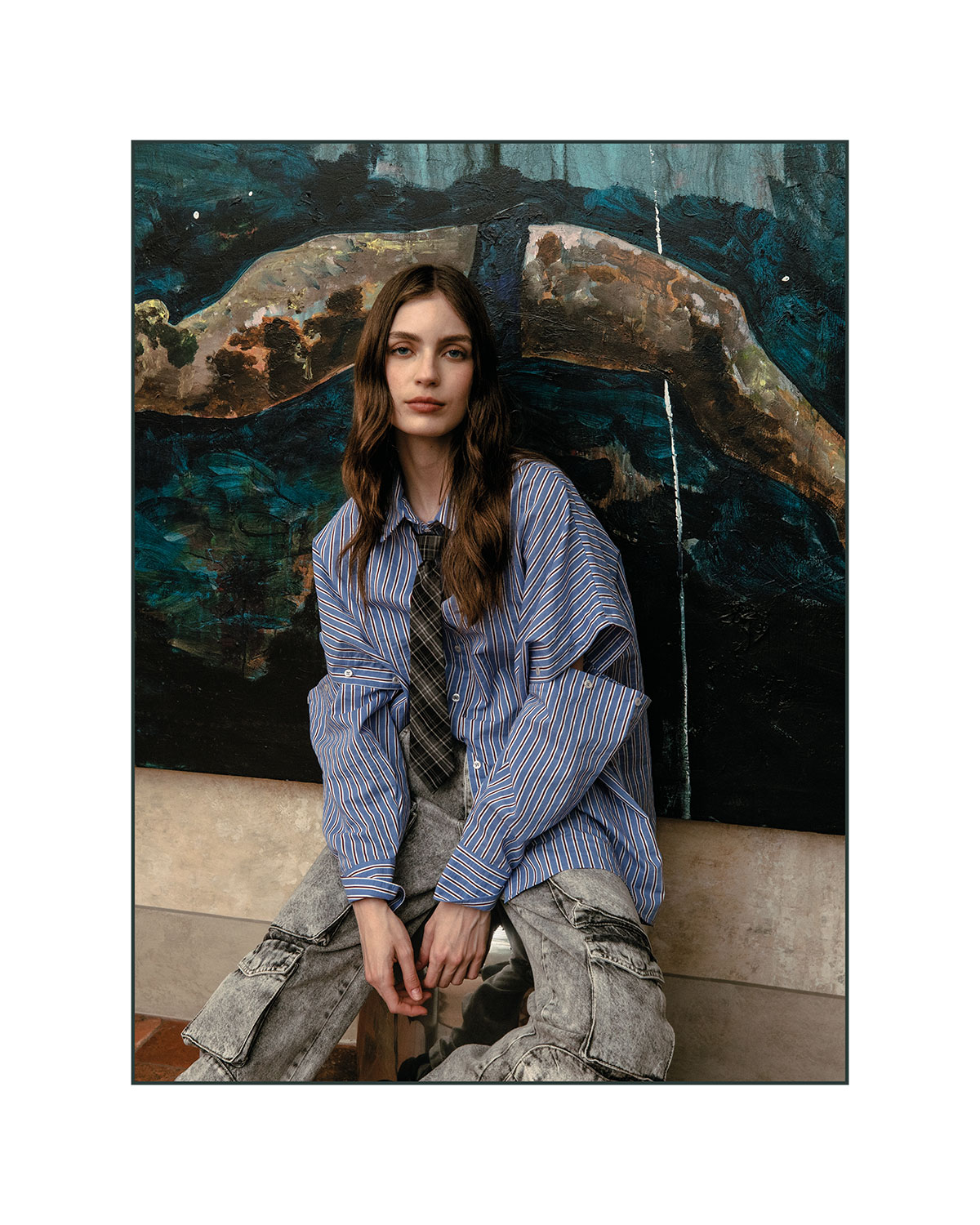 Young woman seated in front of abstract mural wearing oversized blue striped shirt, plaid tie and grey cargo trousers — fashion portrait
