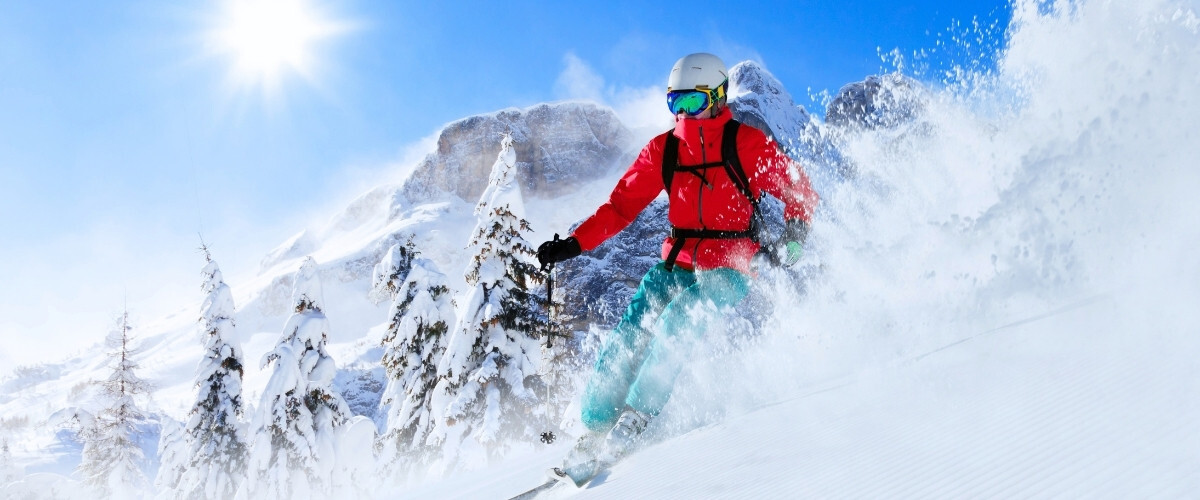 Skier in red jacket and white helmet carving through powder snow on a sunny alpine slope with pine trees