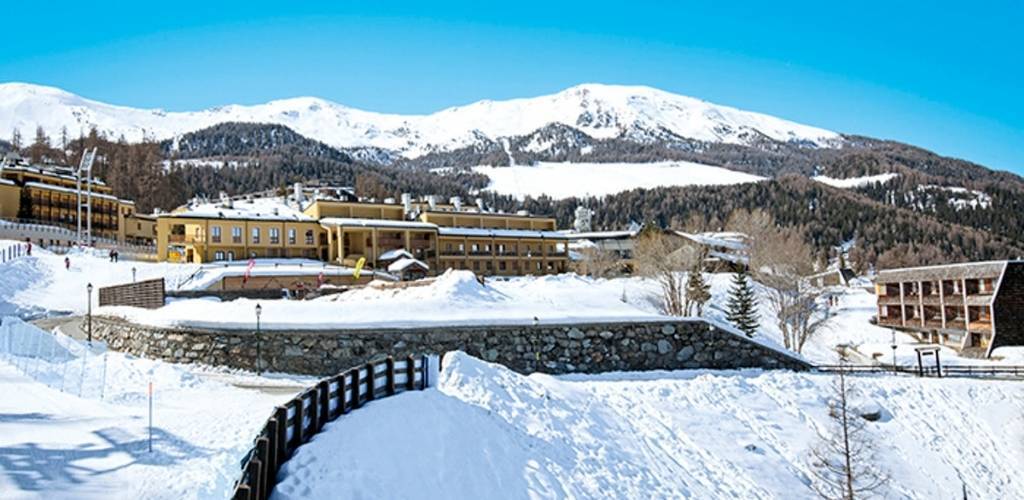Snow-covered alpine resort with yellow lodge buildings, stone terrace, wooden fence and forested mountains under a clear blue sky