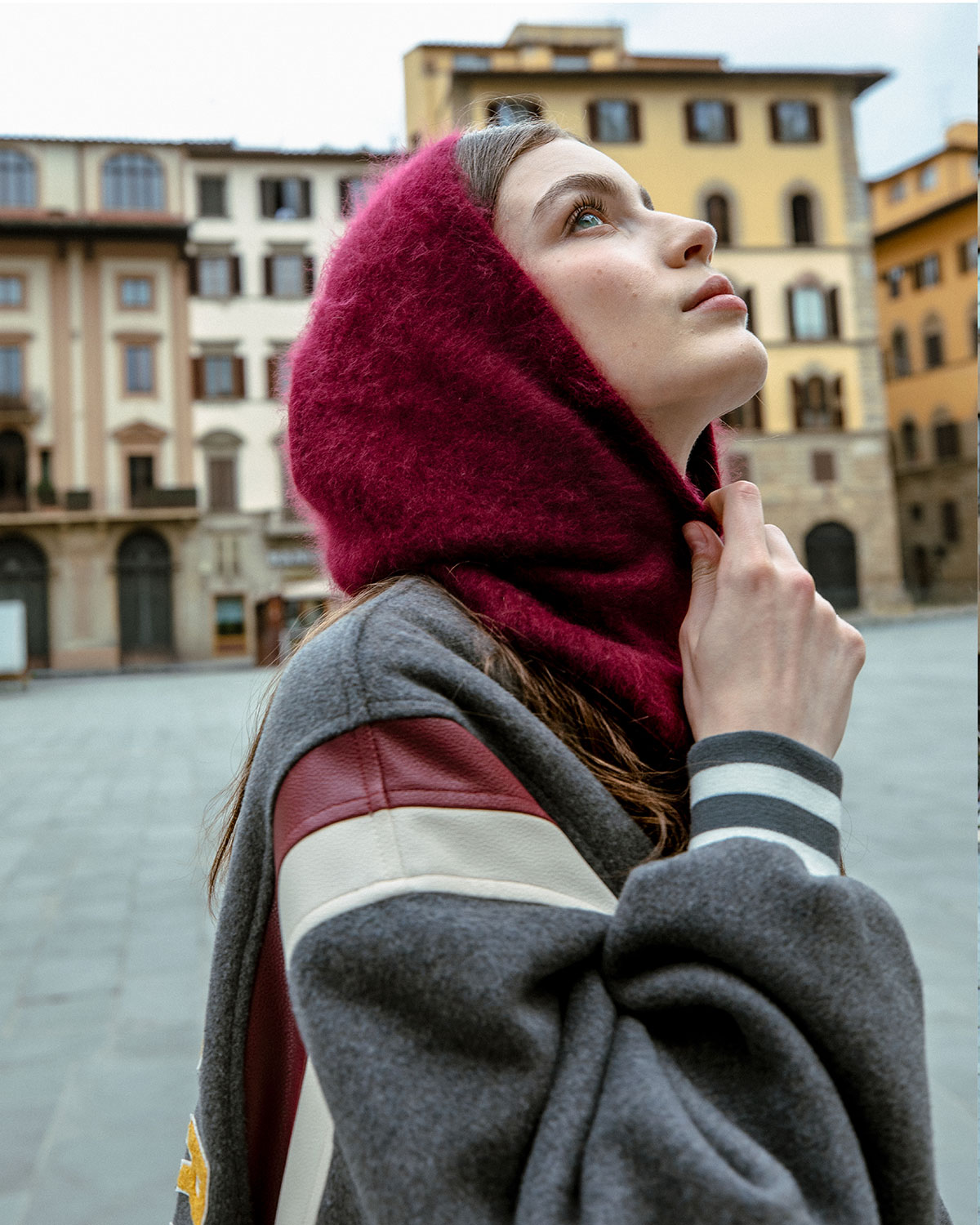 Young woman in a burgundy wool hood looking up in a historic European piazza, wearing grey coat with leather trim