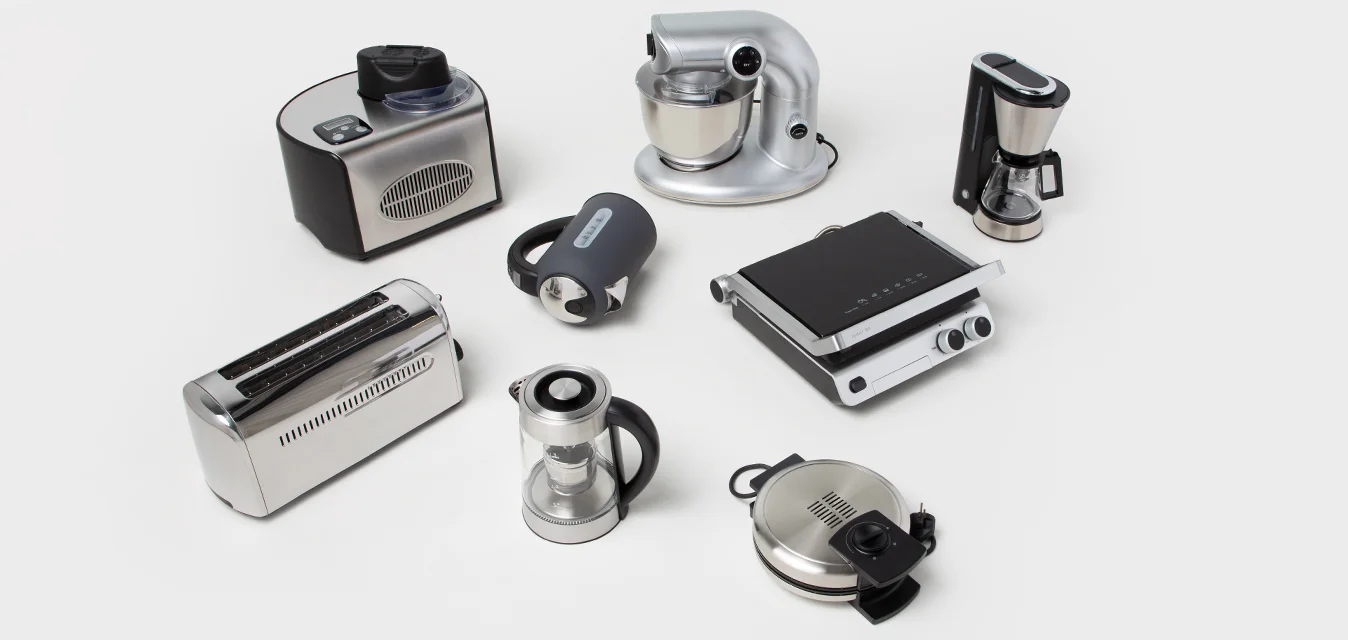 Varie attrezzature da cucina: mixer argento, tostapane nero, caffettiera in vetro, kettle elettrico, griglia elettrica e robot da cucina, tutti con texture diverse.