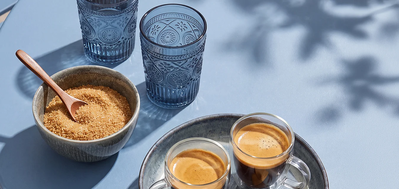 Due tazzine da espresso con biscotti su un vassoio, una ciotola di zucchero di canna con un cucchiaio e due bicchieri d'acqua su un tavolo blu all'aperto.