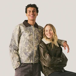 Giovane uomo e donna sorridenti, uomo in piedi con la mano sulla spalla della donna seduta, entrambi indossano giacche casual su sfondo semplice.