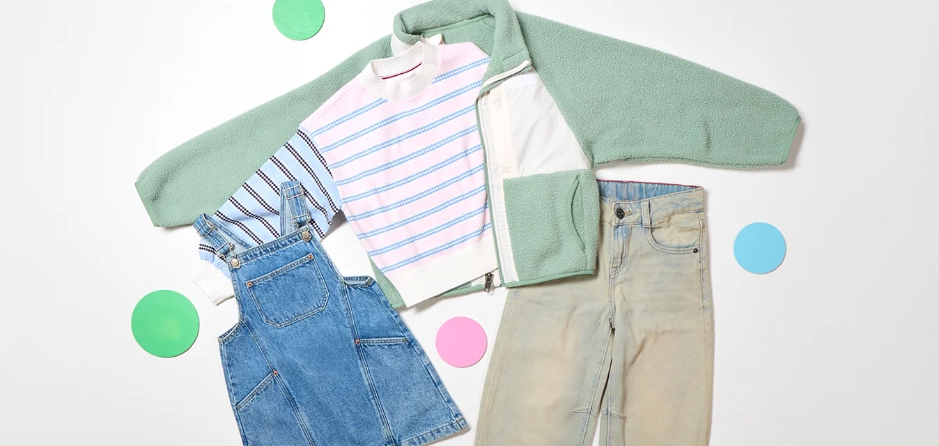 Giacca in pile verde, maglione a righe, abito in denim, pantaloni beige disposti piatti con cerchi di carta colorati su uno sfondo bianco.