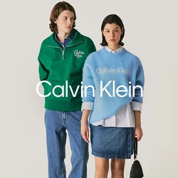 Un uomo in un maglione verde a zip quarti e jeans si trova accanto a una donna in un maglione blu di Calvin Klein, gonna di jeans e che tiene una borsa nera.