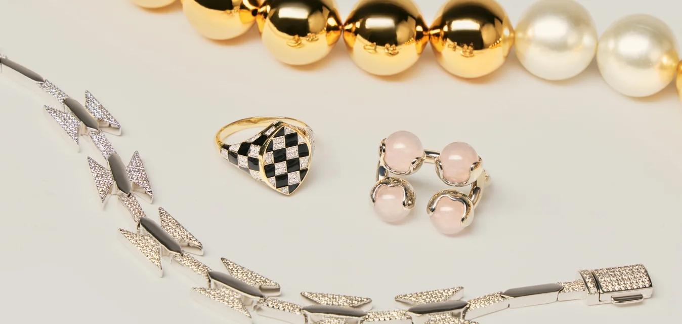 Collana con perline d'oro e perle, anello a scacchi nero e oro, orecchini doppi con pietra rosa e braccialetto geometrico d'argento su superficie bianca.