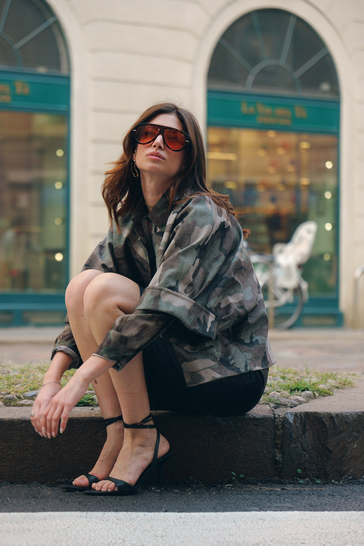 Young woman in sunglasses and camouflage jacket sitting on a city kerb wearing black high-heeled sandals