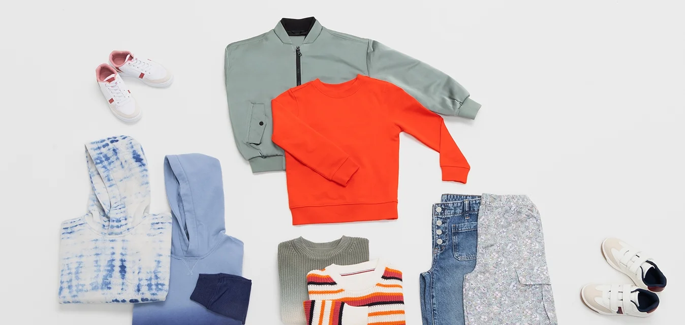 Collezione di abbigliamento: una giacca bomber verde, una felpa arancione, maglioni, jeans in denim con motivo floreale e sneakers bianche e crema.