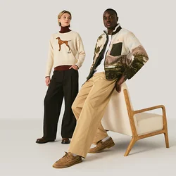 Due modelli posano in uno spazio neutro; uno è in piedi con un maglione con cane e pantaloni neri, l'altro si appoggia a una poltrona di legno indossando una giacca con fantasia e pantaloni beige.