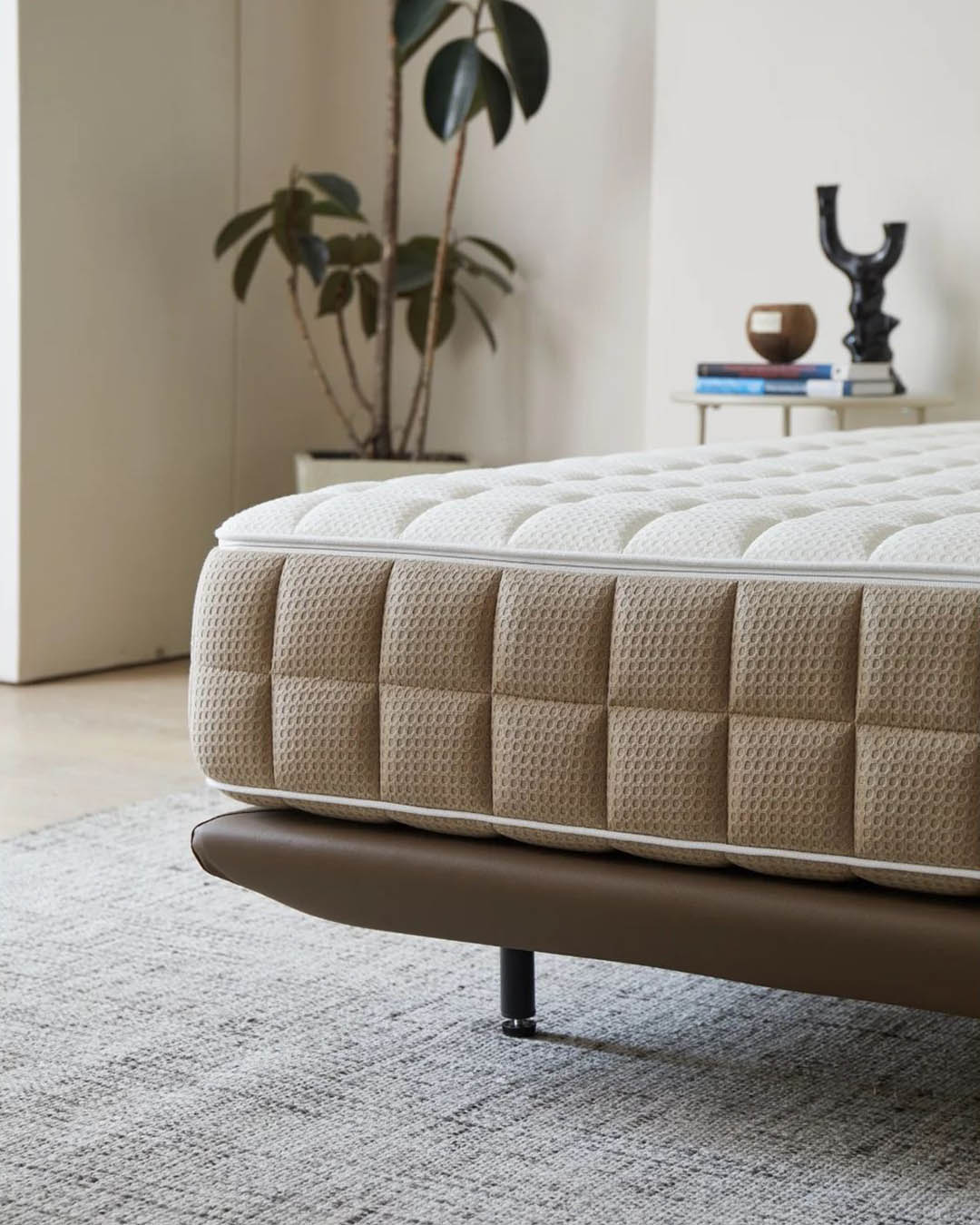 Close-up of a quilted beige and white mattress on a modern brown platform bed frame in a minimalist bedroom with potted plant and bedside table