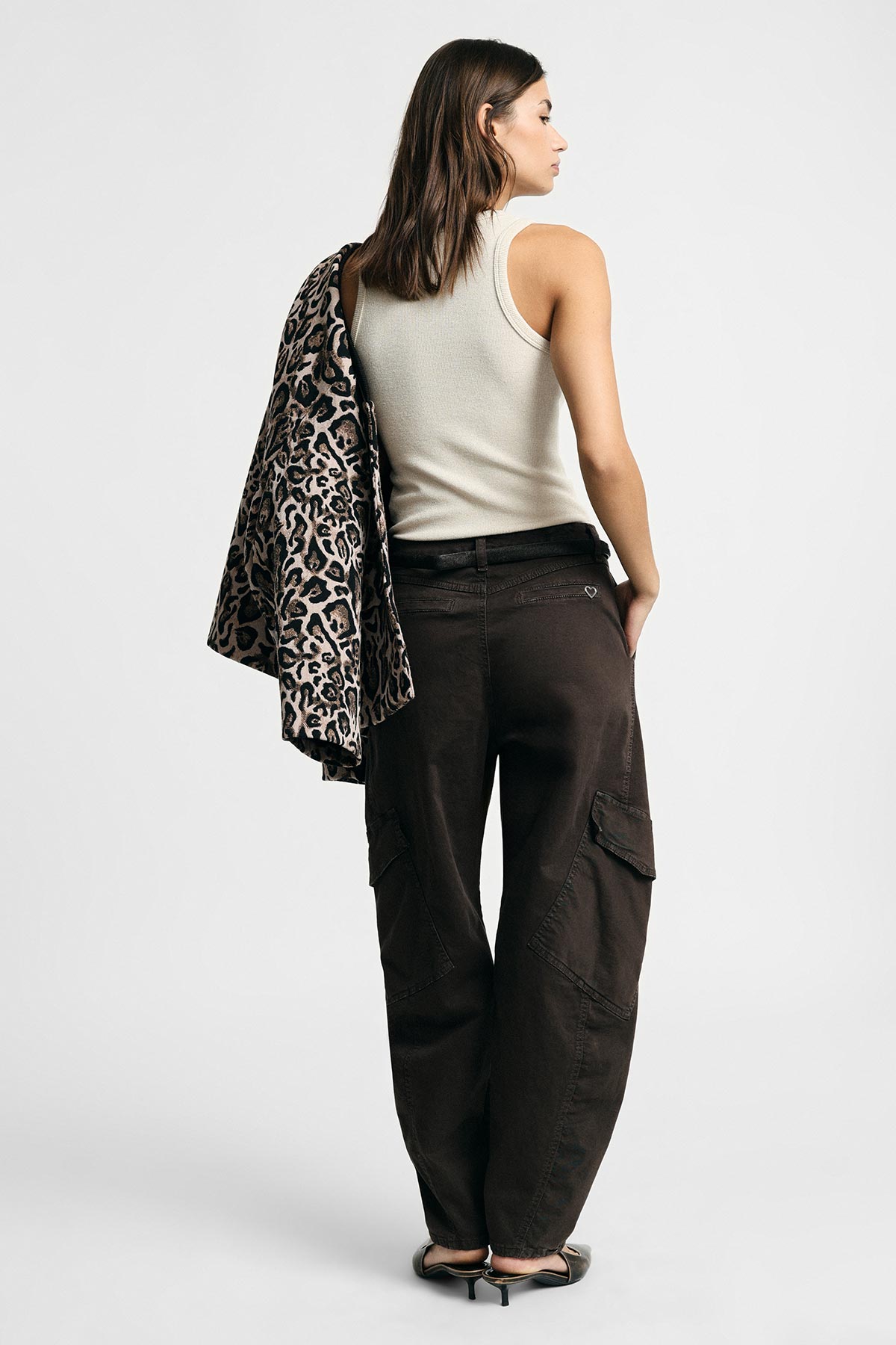 Woman viewed from behind wearing cream tank top, dark cargo trousers and heels, holding leopard-print jacket over shoulder