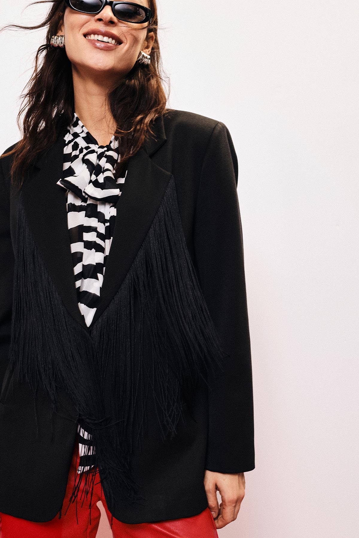 Smiling woman in sunglasses wearing black fringed blazer over zebra-print blouse and red trousers