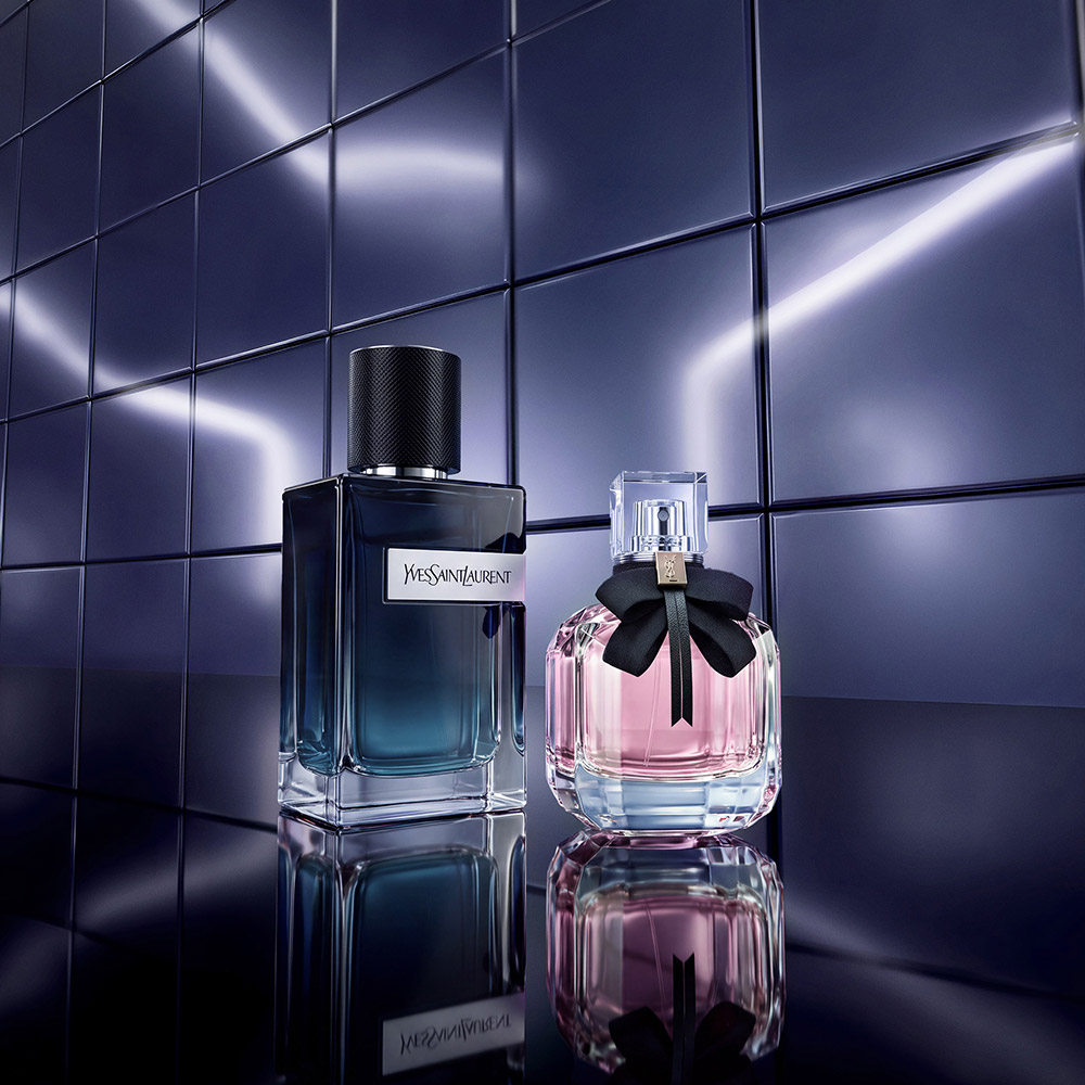I flaconi dei profumi YSL Y Eau de Parfum e Mon Paris esposti su una superficie riflettente lucida davanti a piastrelle blu metallizzato.