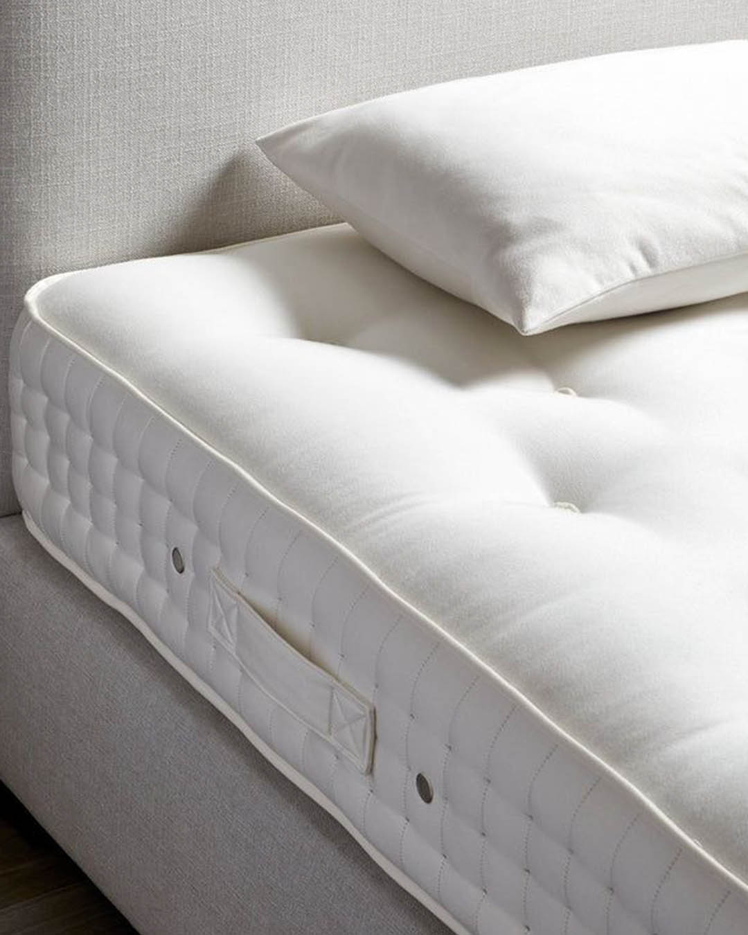 Close-up of a white tufted mattress with side handles and a white pillow on a light upholstered divan