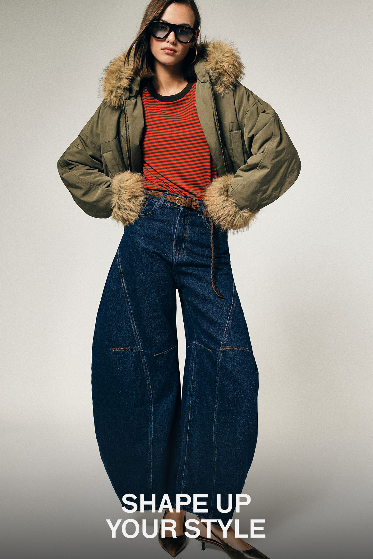 Woman wearing olive fur-trimmed puffer jacket, red striped top and wide-leg denim jeans, hands on hips in studio fashion pose