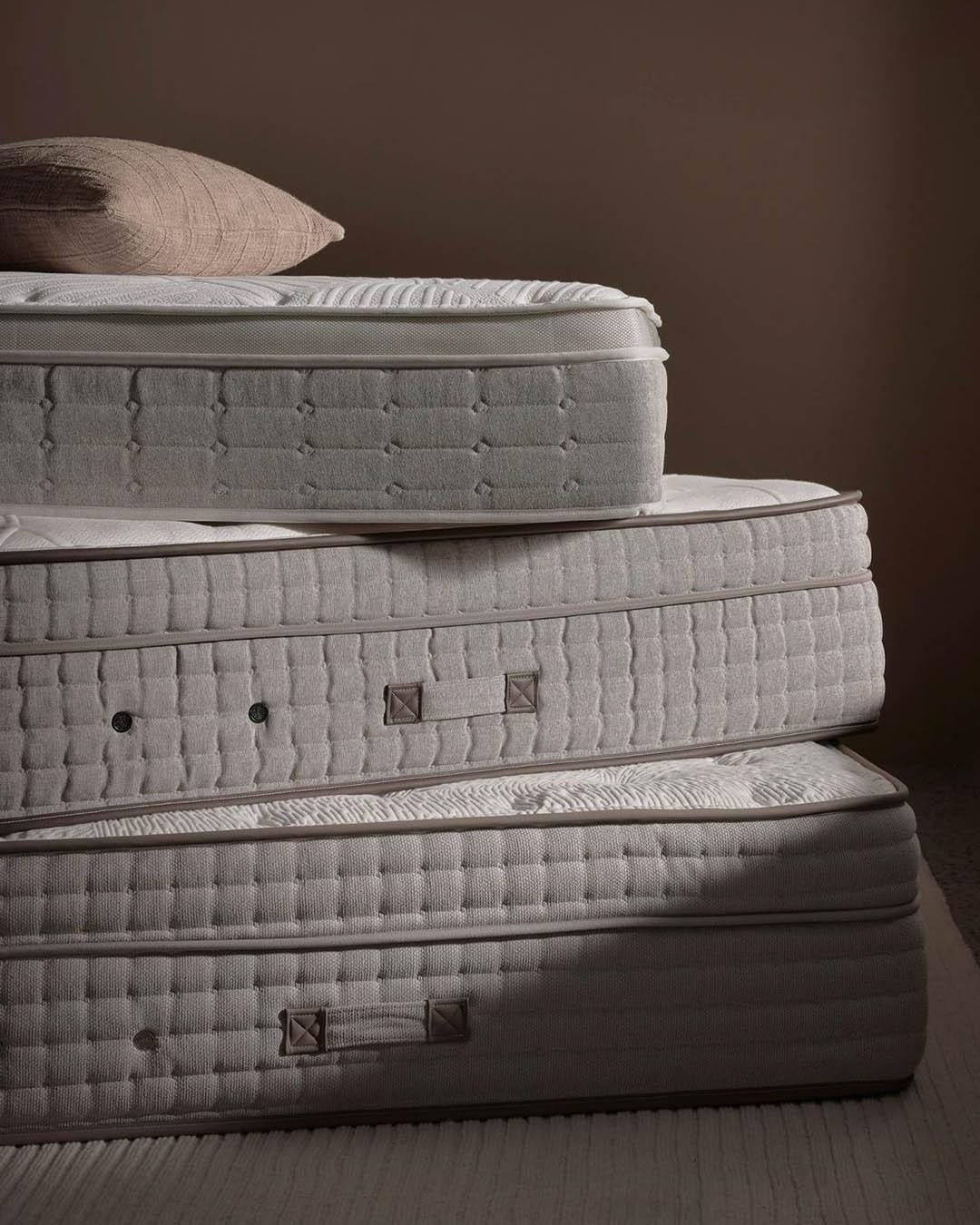 Stack of three plush beige mattresses with quilted fabric, stitched handles and a beige cushion on top against a brown background
