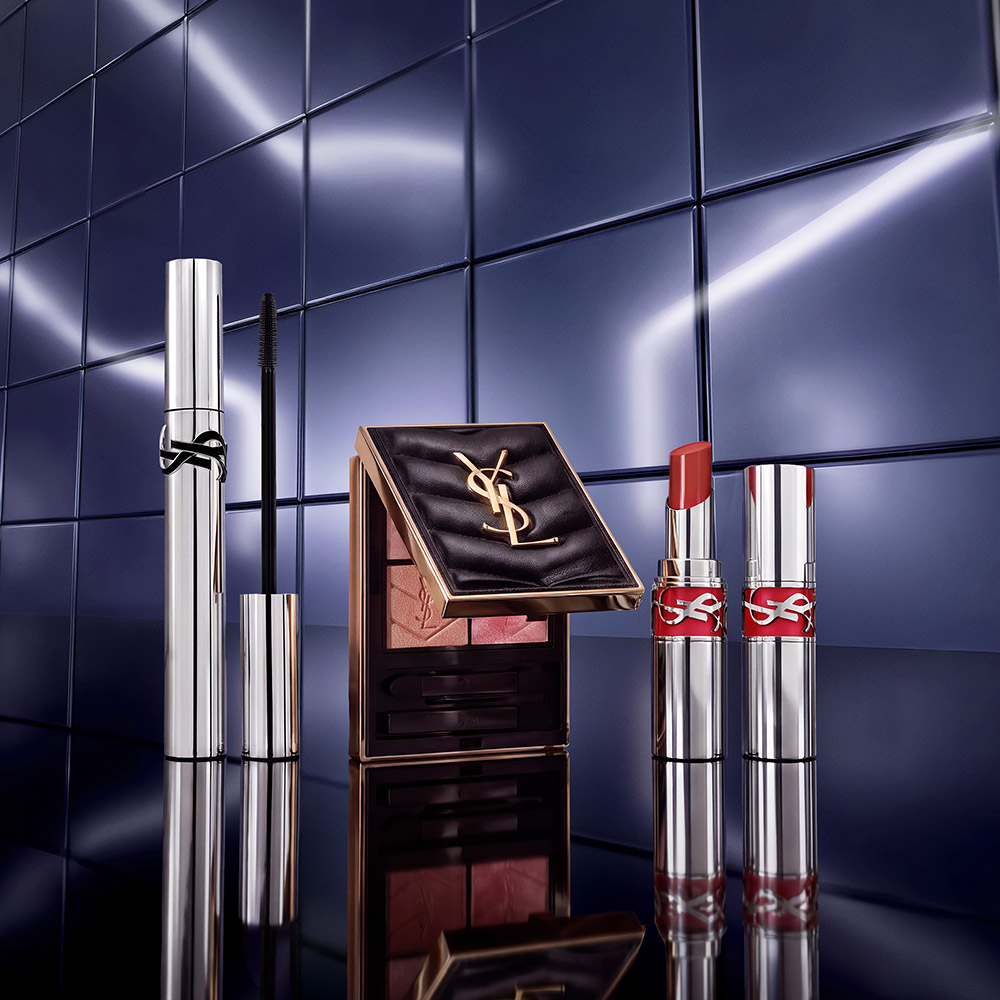 Un mascara argento YSL, una palette di ombretti trapuntata nera e un rossetto corallo su una superficie riflettente davanti a piastrelle blu metallizzato.