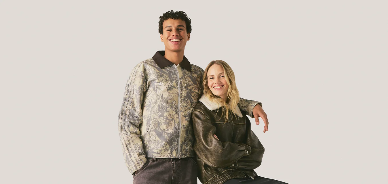 Giovane uomo e donna sorridenti, uomo in piedi con la mano sulla spalla della donna seduta, entrambi indossano giacche casual su sfondo semplice.