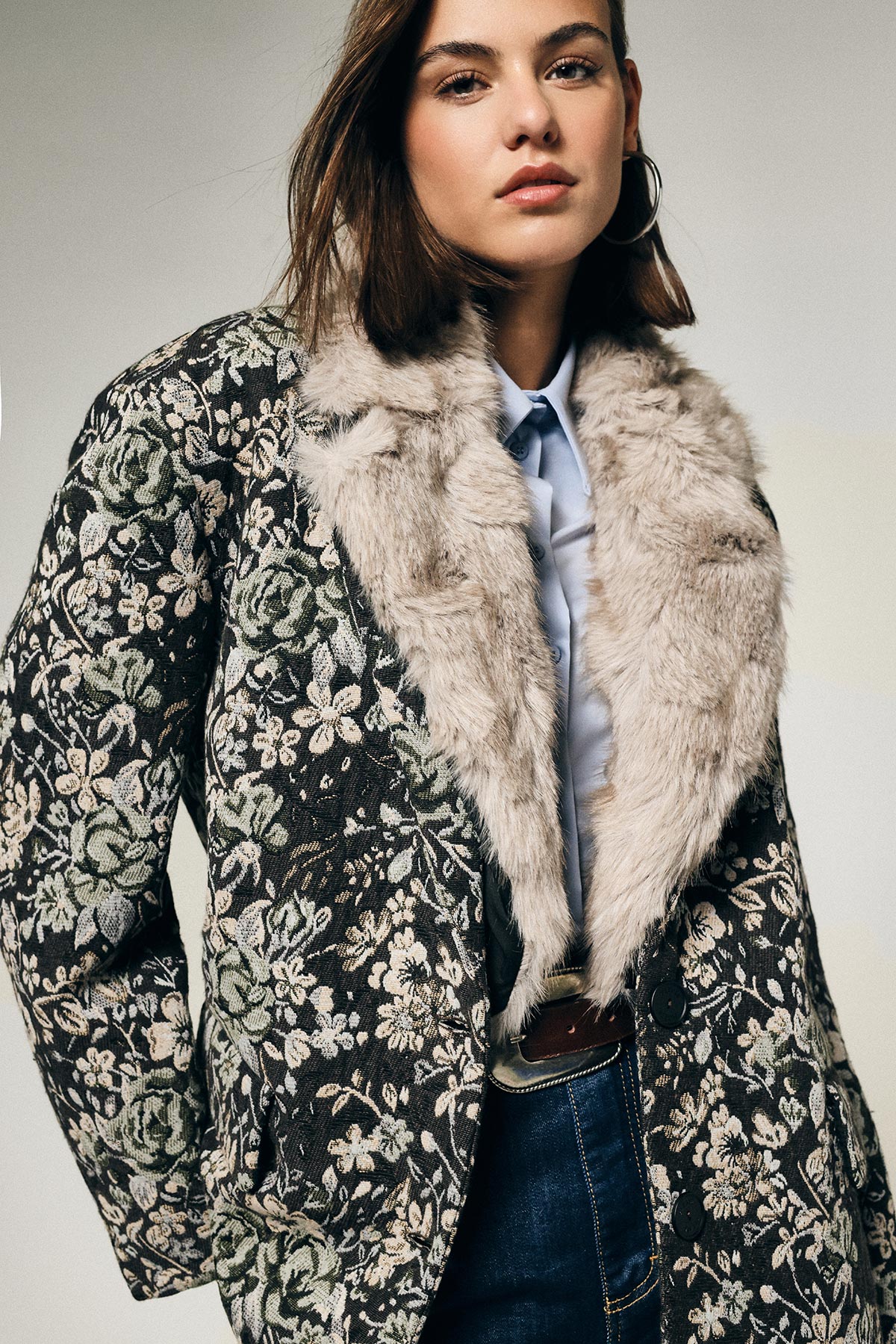 Woman wearing floral brocade coat with faux fur collar, light blue shirt and jeans, hoop earring — fashion portrait