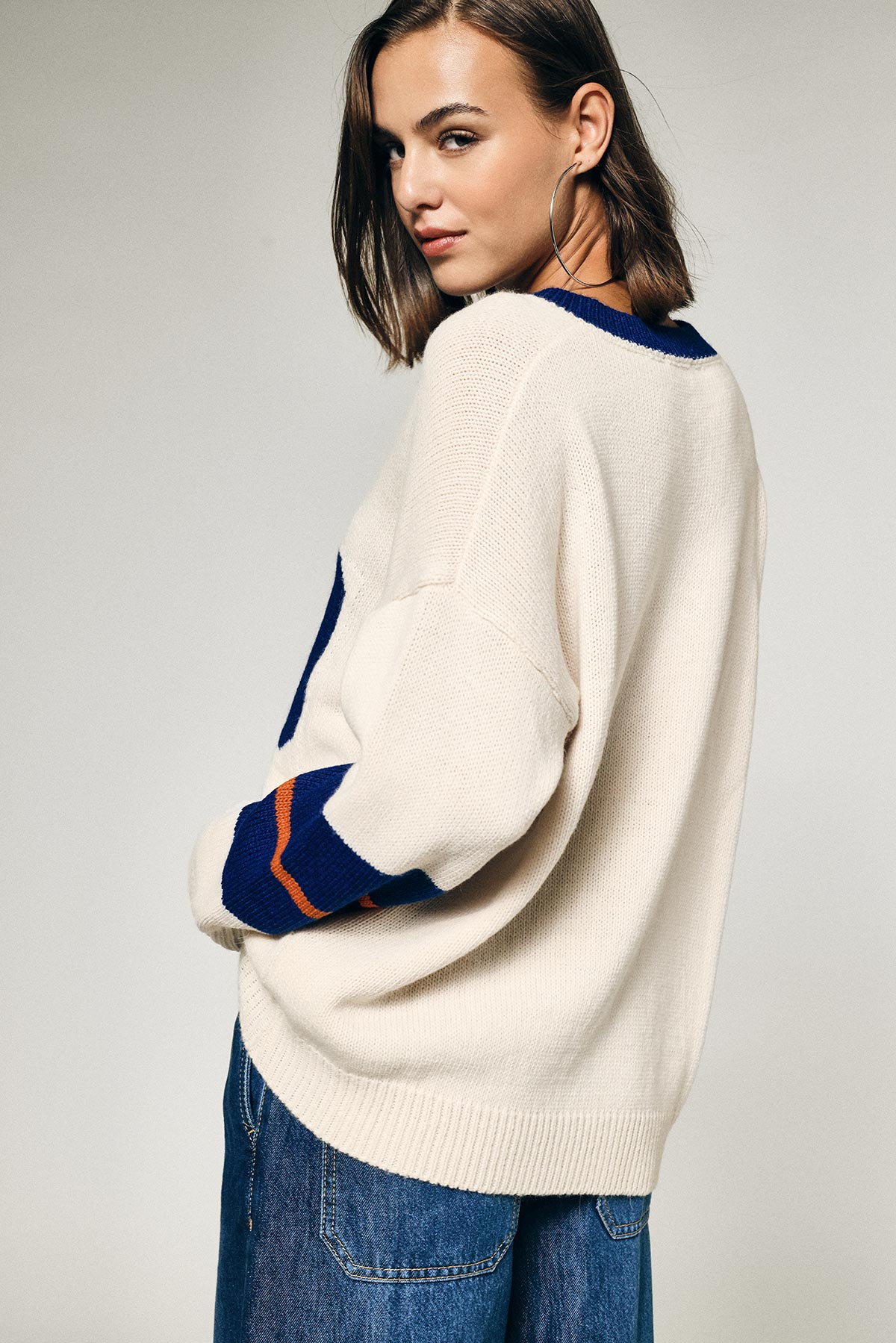 Woman in cream knitted jumper with blue and orange trim and hoop earring, looking over shoulder while wearing denim jeans