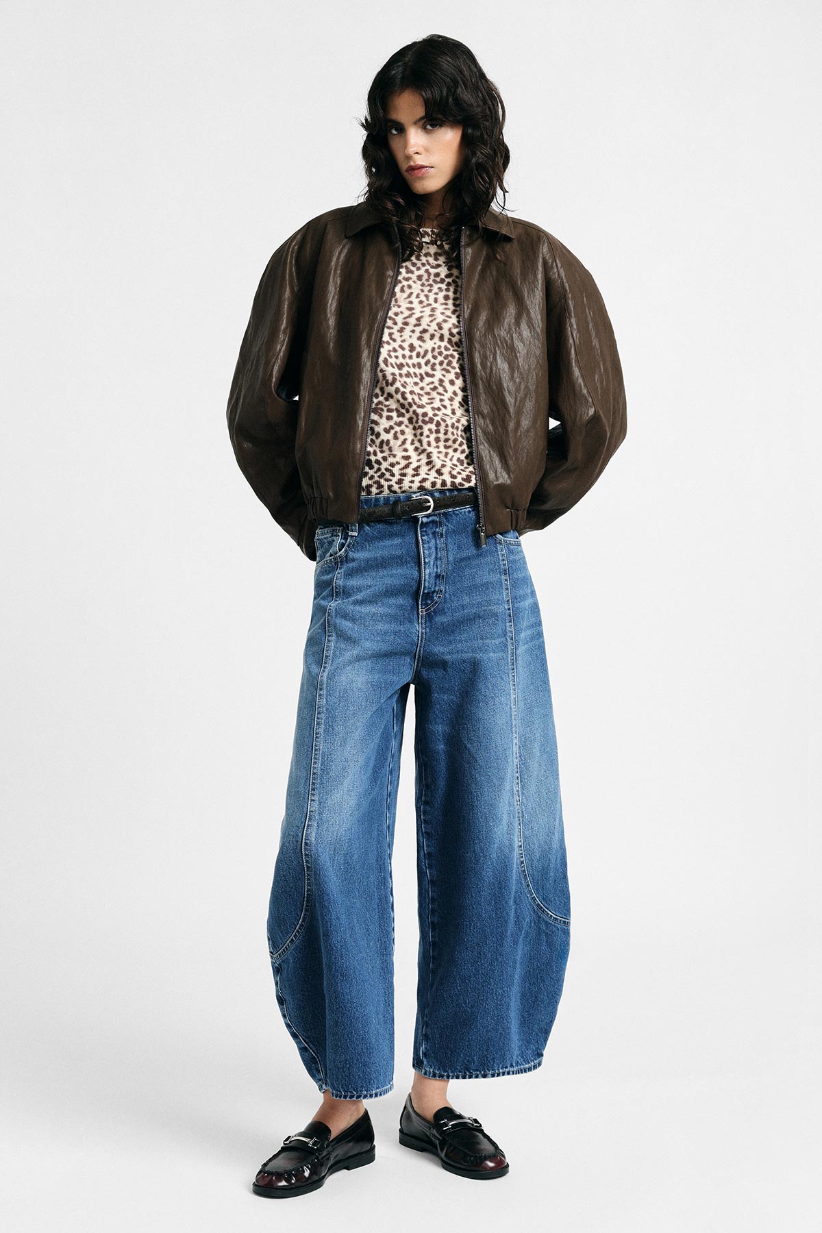 Woman in brown leather bomber jacket, leopard-print top, wide-leg blue jeans and black loafers, white background