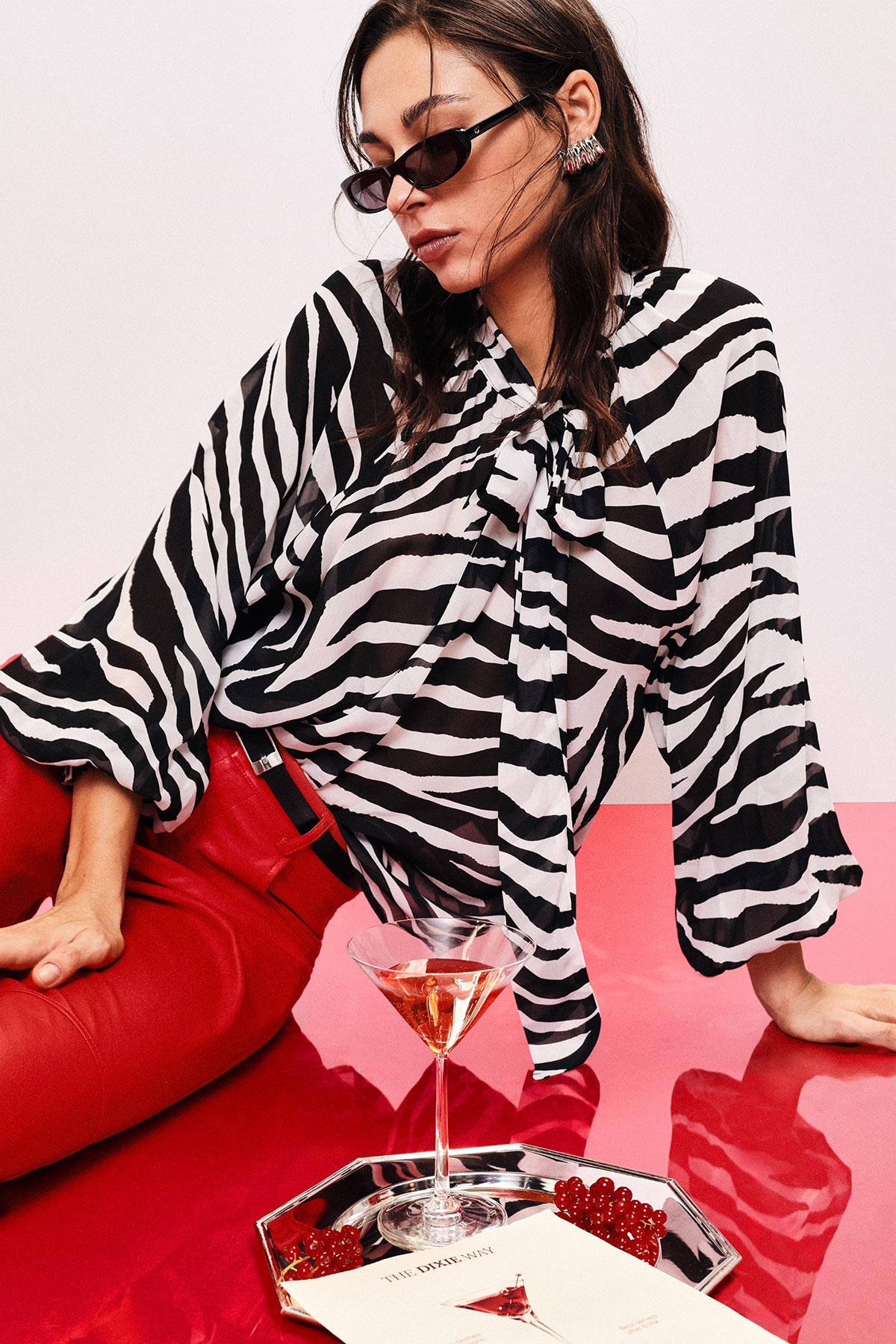 Stylish woman in zebra-print blouse and red trousers wearing sunglasses, leaning on red table with martini glass on tray