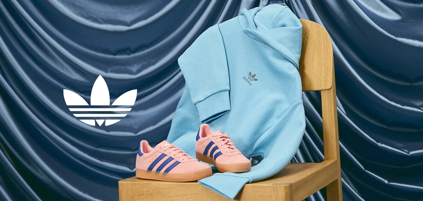 Sneaker Adidas rosa con strisce blu su una sedia di legno, accompagnati da una felpa leggera blu chiaro. Sfondo in tessuto liscio di colore blu scuro.