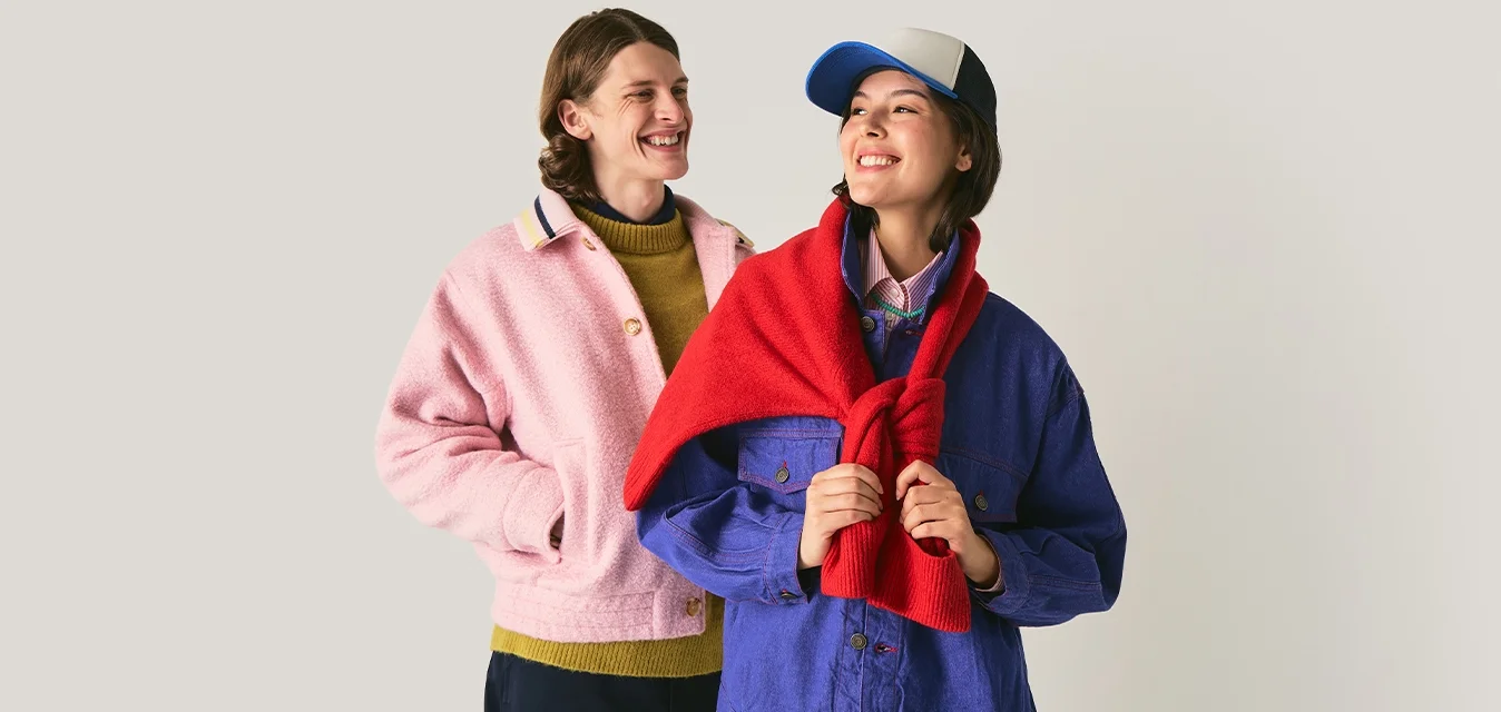 Due persone sorridenti in piedi; una indossa una giacca rosa e un maglione oliva, l'altra indossa una giacca blu, un maglione rosso drappeggiato e un cappellino da baseball.