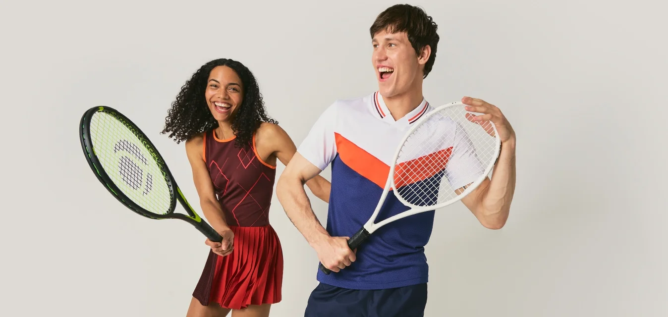 Un uomo e una donna sorridenti in abbigliamento da tennis posano con racchette, in piedi davanti a uno sfondo chiaro e semplice.