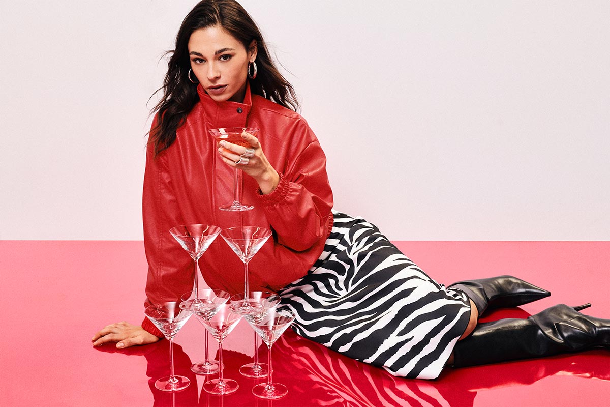 Woman reclining on a red surface in a red jacket and zebra-print skirt, holding a martini glass beside stacked martini glasses and black boots